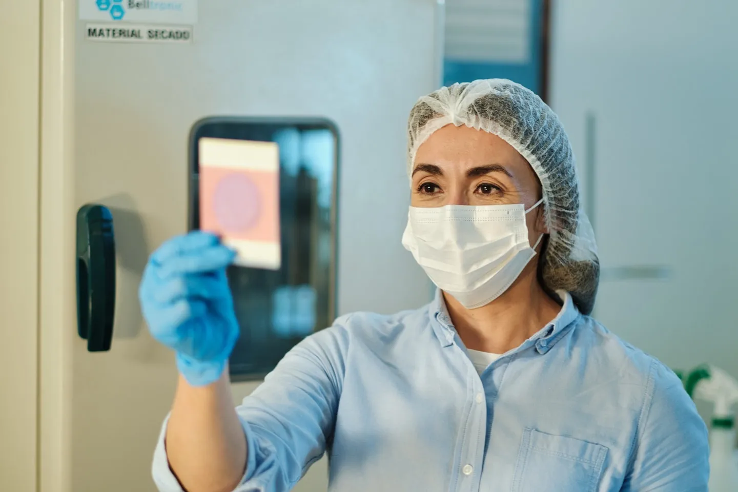 A technician in a hairnet and mask holding a petri dish up to the light